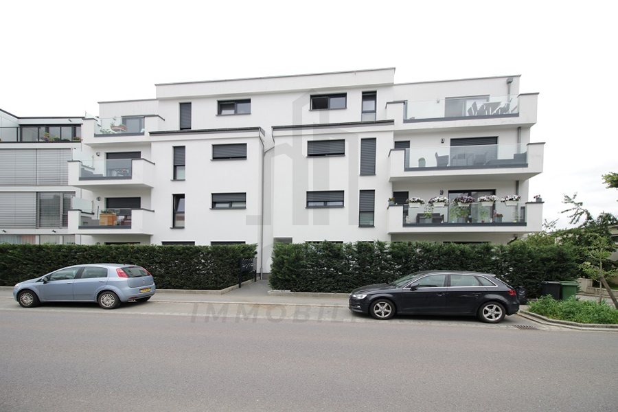 White multi-story apartment residence with dark-framed windows, glass balconies, hedges, and two parked cars along the street.