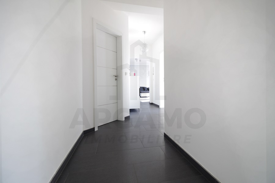 Bright white entrance corridor with dark large-format floor tiles, a closed paneled door, and a glimpse into adjoining rooms.