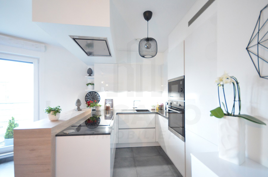 Bright white fitted kitchen with glossy cabinets, built-in appliances, dark counters, a breakfast bar, pendant lights, and an orchid.