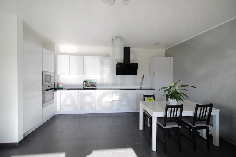 Bright open-plan kitchen and dining area with glossy white fitted cabinets, built-in appliances, dark counters, a black extractor, and a white table.