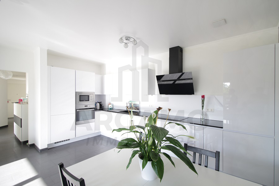 Close view of a glossy white open-plan kitchen with integrated appliances, black worktops, a black extractor hood, and a dining table in front.