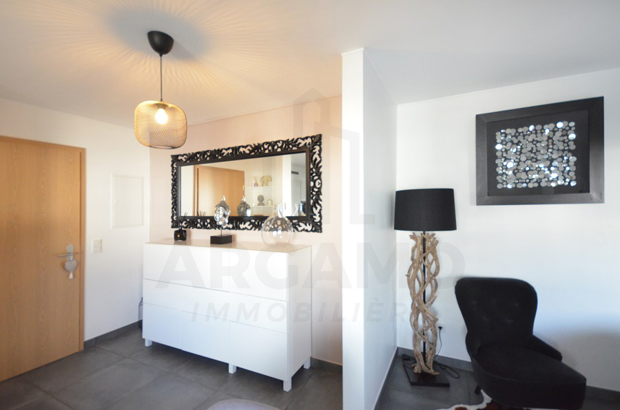 Bright apartment hallway with gray floor tiles, a wooden door, white sideboard, decorative mirror, pendant lamp, black armchair, and floor lamp.