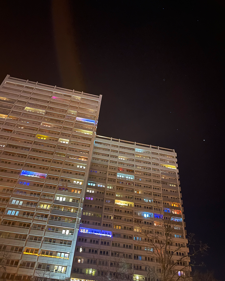Foto eines Plattenblocks mit einzeln bunt beleuchteten Fenstern vor einem dunklen Himmel