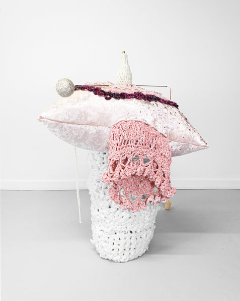 Soft sculpture that is made of mixed materials and a variety of pinks, purples, and whites. A pillow holds a vessel or urn form that sits on top of a crochet while pedestal. On top of the pillow is a pin that has a red thread coming out of it, with a hanging thimble on the end. On the floor is a larger thimble that the smaller thimble hangs into. On the side of the pillow is a loose crochet that looks like it is falling out of the interior of the pillow, and is a darker pink. The form looks bodily.
