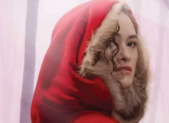 Against a white misty background, a fair-skinned woman's face is bordered by a blood-red hood with fur trim.