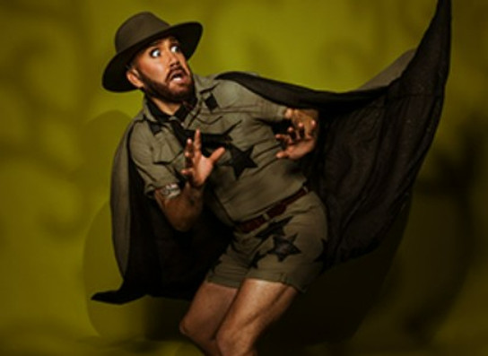 A tan-skinned amn with a beard pulls a fearful expression, looking over his shoulder with his handsraised in alarm. He wears a khaki 'Scouts'-styled uniform, and a long olie-green cape billows behind him. He has an akubra-styled hat on.