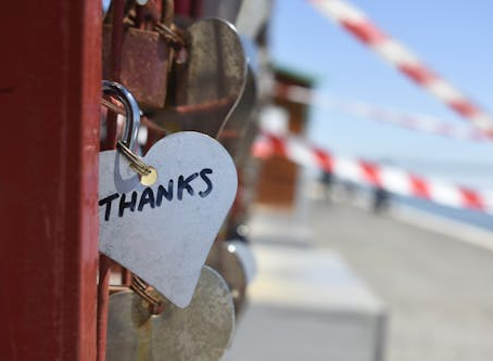 A close-up of heart-shaped padlocks attached to a red railing, with one silver heart lock engraved with the word “Thanks,” and a blurred walkway and striped barrier in the background.