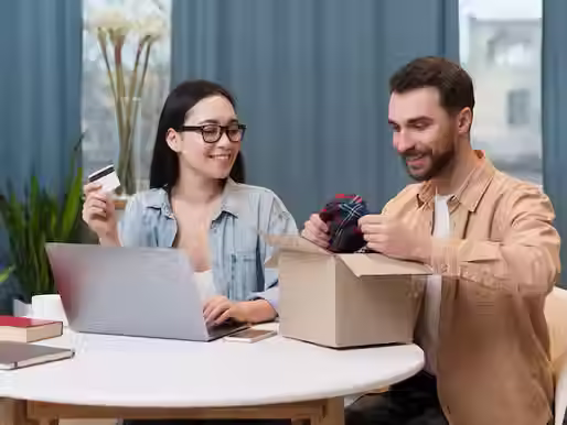 A happy couple unboxing an online purchase at home. The woman, wearing glasses and a denim shirt, holds a credit card while sitting in front of a laptop. The man, wearing a beige jacket, smiles as he takes a plaid fabric item out of a cardboard box. A cozy indoor setting with books and a cup on the table.
