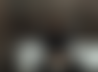 A man sits on a narrow bed in a dim, bare room, leaning forward with his hands clasped and head bowed, conveying a somber and reflective mood.