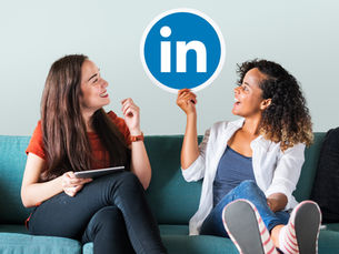 Two cheerful women sitting on a couch, holding a LinkedIn logo cutout and discussing professional networking, symbolizing social media marketing and LinkedIn engagement strategy.