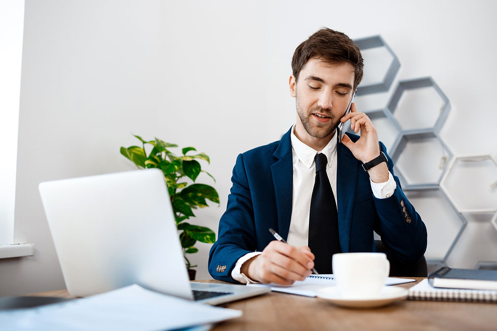 Business professional speaking on a smartphone while taking notes at a desk with a laptop in a modern office.