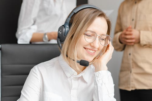 A smiling young woman wearing glasses and a headset with a microphone, sitting at a desk in an office. She appears engaged in a conversation or call, with two colleagues blurred in the background.