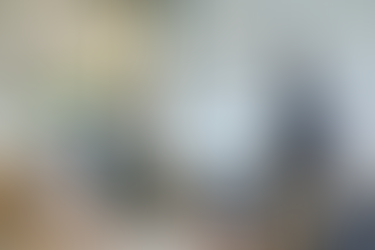 Four colleagues wearing face masks meet in a modern office conference room. A man stands beside a screen displaying pie charts and the heading “Sales: Find your customers,” while a woman holding papers and two seated coworkers listen and look toward the presentation.