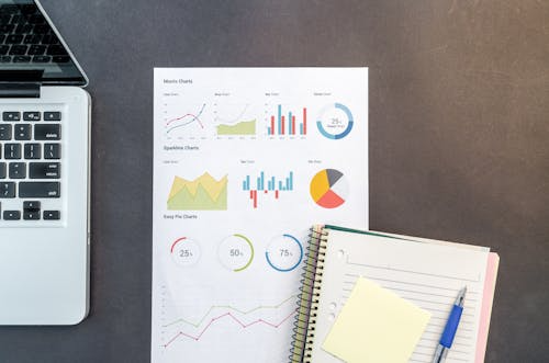 Overhead view of a workspace with a laptop, printed analytics charts showing graphs and percentages, a spiral notebook, pen, and sticky notes, representing data analysis and performance tracking.