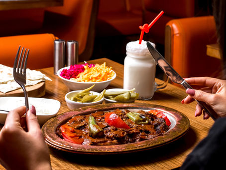 Person dining at a restaurant with a plate of grilled meat and vegetables, served with pickles, salad, and a yogurt drink.