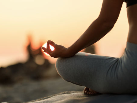 Close-up of a woman sitting in a lotus meditation pose at sunset, her hand resting on her knee in a relaxed mudra, with warm golden light and a softly blurred natural background creating a calm, mindful atmosphere.