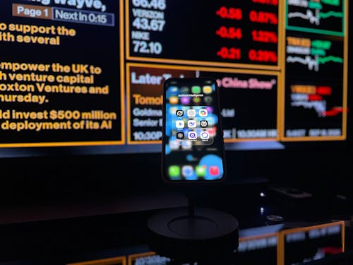 A smartphone mounted on a stand in front of a large digital financial news and market data screen, showing app icons on the phone display with stock prices, headlines, and red and green market indicators in the background.