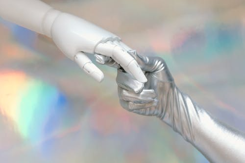 Close-up image of a white robotic hand gently touching a silver, human-like gloved hand against a soft, iridescent background, symbolizing the connection between artificial intelligence and human interaction.
