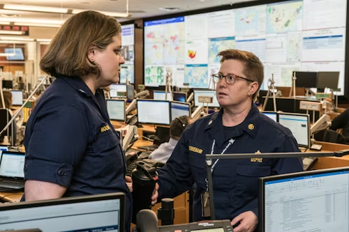 Two uniformed professionals stand in a busy operations control room, engaged in a focused conversation. Multiple computer monitors and large wall screens display maps, data dashboards, and system information in the background. One person gestures while speaking, suggesting coordination or decision-making in a high-pressure, data-driven environment.