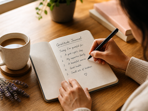 Person writing in a gratitude journal at a warm wooden desk with tea, books, and soft natural light, creating a calm and reflective journaling space.