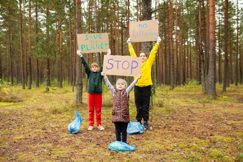 Three children stand in a forest holding signs that read "SAVE THE PLANET," "no Littering," and "STOP," with blue garbage bags on the ground around them.