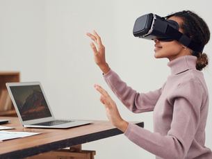 A woman wearing a virtual reality headset sits at a wooden desk, interacting with the air using her hands. An open laptop and documents are on the table beside her, suggesting she is engaged in a virtual simulation or remote work environment.