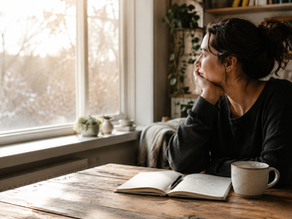 Person sitting at a wooden table beside a sunlit window, reflecting quietly with an open journal, pen, and cup of tea in a cozy home setting