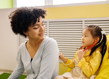 A woman smiling by looking at a little girl wearing yellow dress and holding a toy stethoscope pretending to do the checkup of the lady.