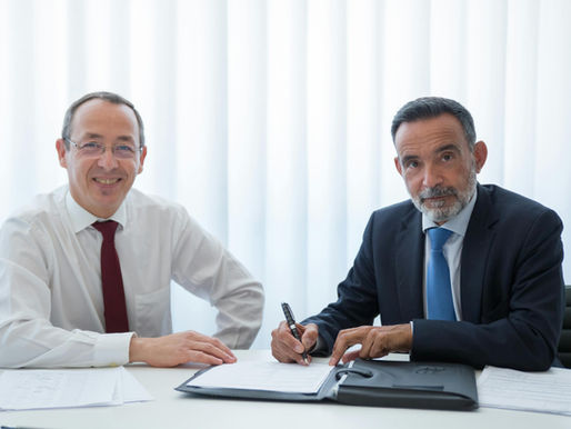 Two professional businessmen sitting at a desk in a bright office environment. One man is smiling at the camera while the other is signing a document, symbolizing a formal business agreement. The scene represents a successful B2B enterprise sales meeting or partnership.