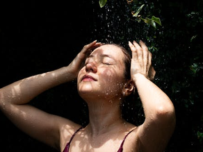 Woman with eyes closed enjoying falling water, hands in her hair, standing outdoors against a dark leafy background, conveying calm and refreshment.