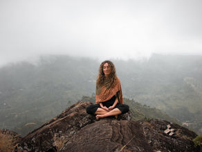 Woman meditating in a cross-legged position on a rocky mountaintop, surrounded by misty mountains in the background. She is wearing a patterned shawl and black pants, with her sandals placed beside her on the rock.