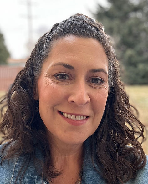 Photo of Desta Jones. She has long, curly brown hair, is 52 years old, and is smiling. She is sitting outside in the sunlight and is surrounded by trees.