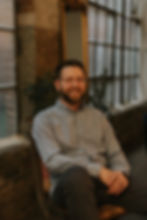 Smiling Perinatal Mental Health Specialist and Therapist in glasses sits in a chair, wearing a patterned shirt. Background shows industrial windows and a plant. Warm indoor setting.