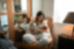 Mother in striped pajamas sits in a chair, cradling a baby and holding a mug. Soft light filters through a window, creating a cozy mood.