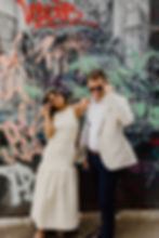 A bride in a white sleeveless dress and a groom in black pants, white shirt and a cream jacket are posing in front of a wall painted with lots of graffiti. The are both pulling their sunglasses down from their eyes.