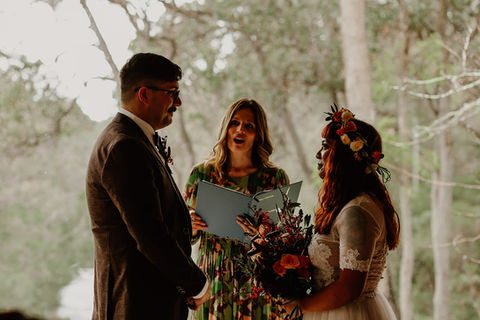 Three people from the waist up during an elopement marriage ceremony. On the left, the groom, a man in a brown suit and glasses in profile. In the middle, the celebrant faces the front, long light brown wavy hair, she holds a blue folder. On the right, the bride in a lacy short sleeved dress holds a bright coloured bouquet of flowers that match the flower crown in her long red hair, is in profile.