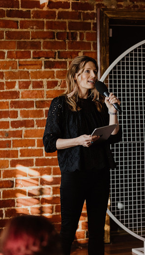 Sally Bruce Celebrant in black pants and a loose black top holds a microphone and an iPad in front of an exposed brick wall at an urban Perth wedding.