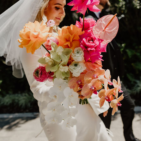 Bride in a white dress and veil holds vibrant bouquet of pink, orange, and white flowers outdoors. Pathway and greenery in background.