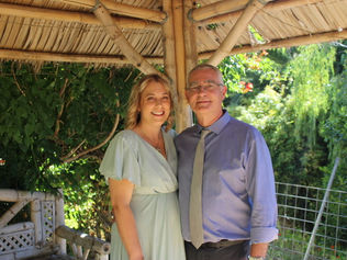 A woman in a light dress and a man in a blue shirt smile under a bamboo gazebo. Lush greenery surrounds them on a sunny day. They've just eloped in the Perth Hills with Sally Bruce Celebrant