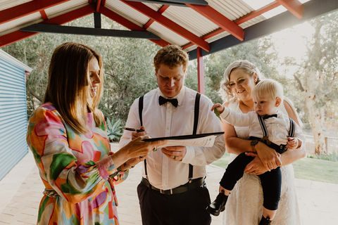 Female marriage celebrant in brightly coloured dress is holding a folder while a man in a white shirt, blue bow tie and suspenders signs the paperwork. Watching next to the man is a blonde-haired bride in a white dress holding a toddler who is in matching shirt, bow tie and suspenders.