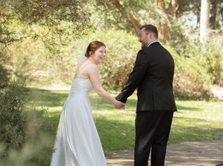 Bride in white dress and groom in black suit hold hands, smiling in a sunlit garden with green foliage and path in the background. It's a Kings Park elopement with Sally Bruce Celebrant.