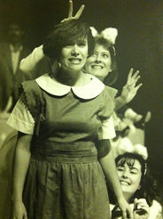 Black and white photo - a woman with dark hair in a dress with a large white collar stands in front of two women dressed as cats; one above her who holds rabbit ears over her head, and one below her. The cats are smiling; the woman looks frustrated.