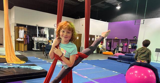 Young girl smiles while hanging from red aerial silks in gymnasium