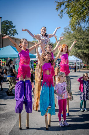 Young girls standing on stilts, arms raised in a group, event outdoors.