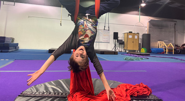 Young person hanging upside down on aerial silks smiling happily for the camera.