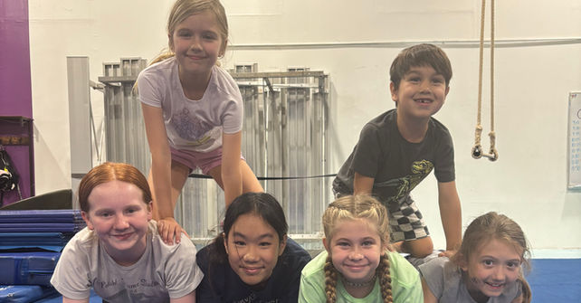 Gymnastics class children stacked, smiling, forming human pyramid, on blue mat inside gym.