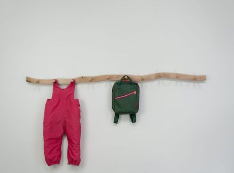 a pair of pink overalls and a green backpack are hanging on a wooden coat rack