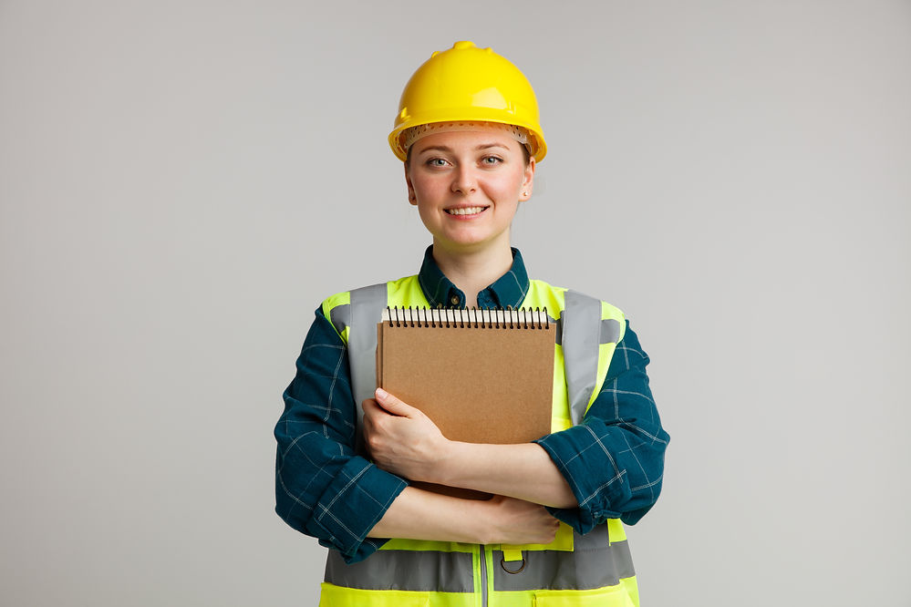 smiling-young-female-construction-worker-wearing-safety-helmet-safety-vest-hugging-notepad