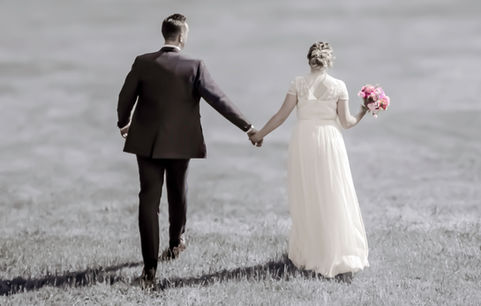 A black and white rear image of the bride and groom walking to the reception with the bouquet detailed in colour