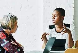 Two women seated at a table having a discussion, with one using a laptop while speaking.